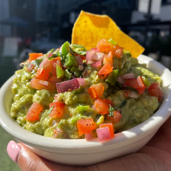 A hand holds a white bowl of guacamole topped with diced tomatoes, red onions, and a tortilla chip.