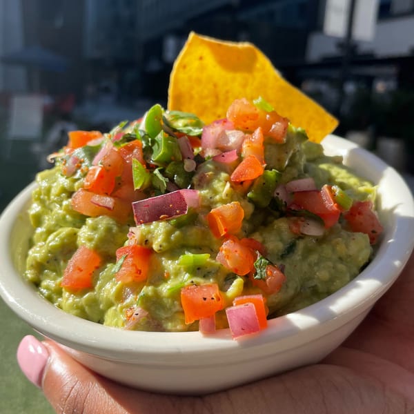 A hand holds a white bowl of guacamole topped with diced tomatoes, red onions, and a tortilla chip.