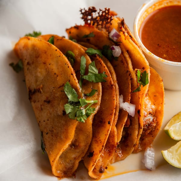 Five Pretty Little Tacos with cilantro, onions, lime wedges, and a side of birria consommé.